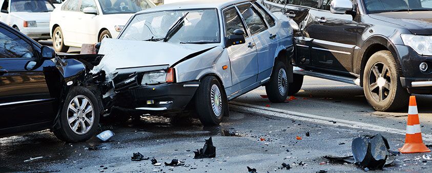 Three cars involved in a collision on a city street, with visible front-end damage, debris scattered on the road, and an orange traffic cone nearby.