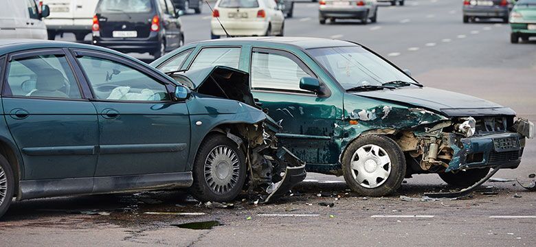 Two cars with severe front-end and side damage are stopped in the middle of a busy road after a collision, with other vehicles visible in the background.