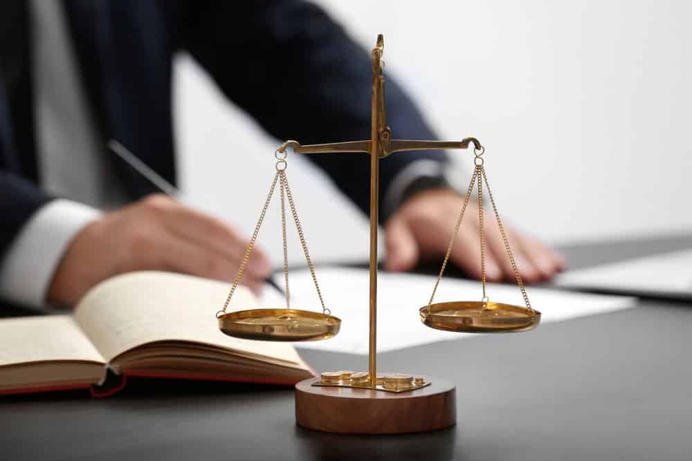 A close-up of golden balance scales on a desk with an open notebook. In the background, a person in a suit is writing on paper, suggesting a legal or judicial setting.