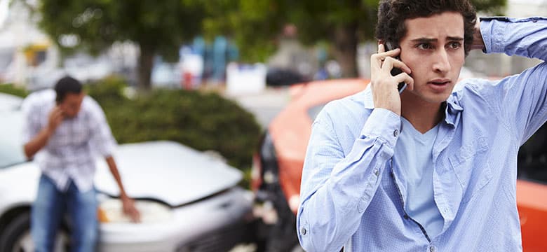 A young man talks on his phone with a worried expression, standing in front of two cars involved in an accident. Another man is in the background also making a phone call, with the damaged cars behind them.