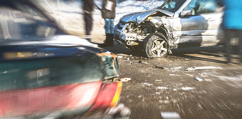 Two cars after a collision on a road; one car has severe front-end damage, while the other is partially visible. Debris is scattered on the ground, and several blurred people are standing nearby.