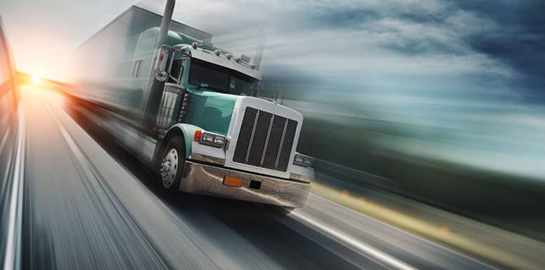A large green semi-truck speeds down a highway at sunset, with motion blur emphasizing its fast movement. The sky is partly cloudy and the road stretches into the distance.