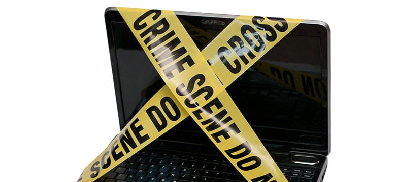 A laptop with its screen and keyboard covered by yellow Crime Scene Do Not Cross tape, suggesting a digital or cyber crime investigation.