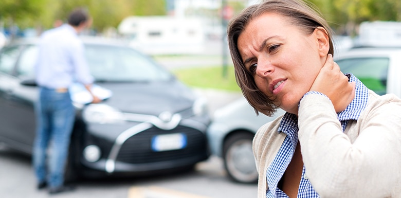 A woman holding her neck in pain stands in front of two cars that appear to have been in an accident. In the background, a man is inspecting the vehicles and writing on a clipboard.