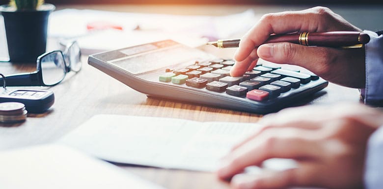 Close-up of a person’s hands using a calculator and holding a pen on a wooden desk, with documents, glasses, and coins nearby, suggesting financial or accounting work.