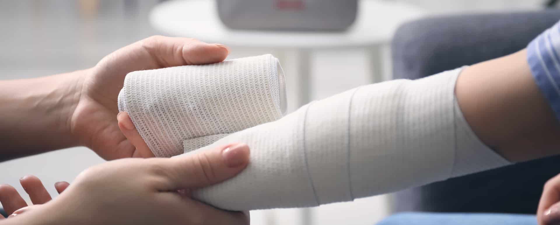 Close-up of a person wrapping another person’s forearm with a white bandage, suggesting care for an injury, with a blurred background showing a table and chair.