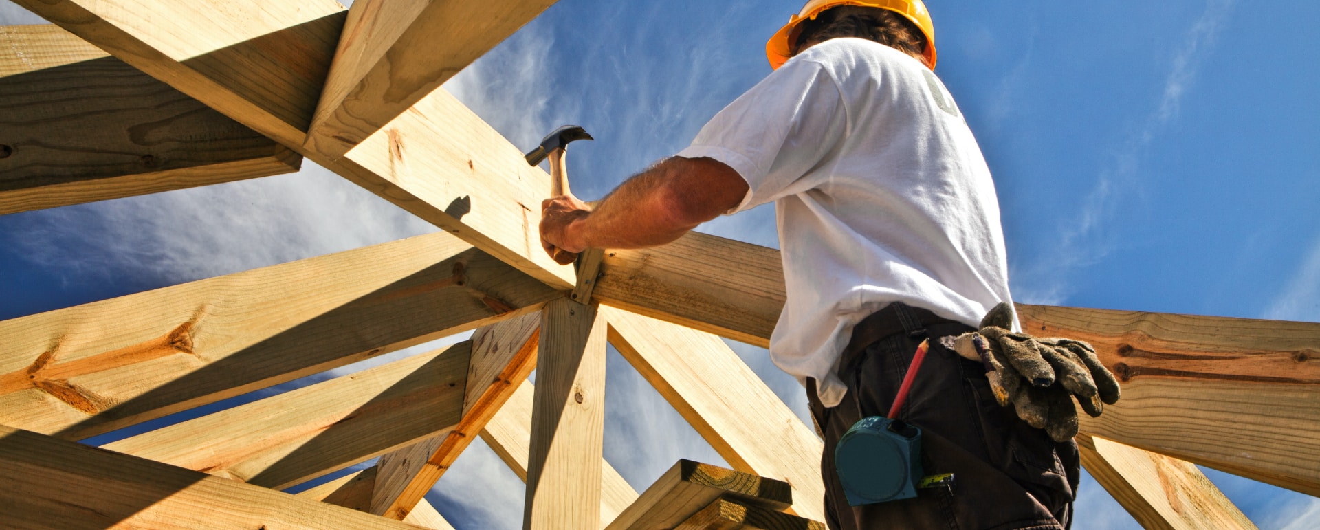 A construction worker wearing a hard hat uses a hammer to secure wooden beams while building a roof frame under a blue sky. Gloves and a measuring tape hang from his belt.