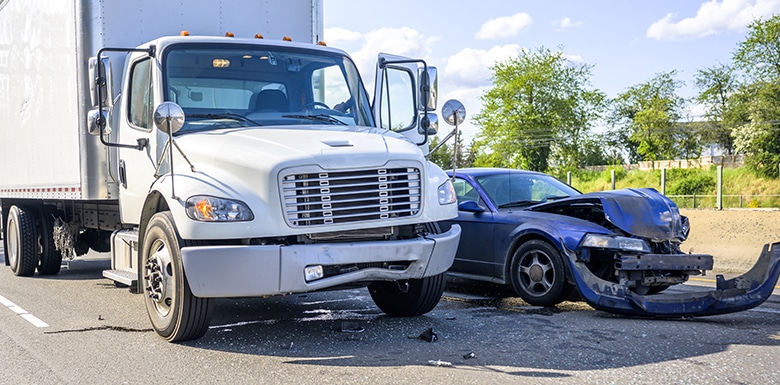 A white box truck and a blue car involved in a collision on a road. The front of the car is severely damaged, with its bumper detached, while the truck has minor visible damage. Debris is scattered on the road.