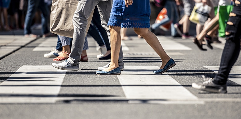 Close-up of several people’s legs as they walk across a crosswalk on a city street, with some carrying shopping bags. The scene is sunny and the focus is on the lower half of their bodies.