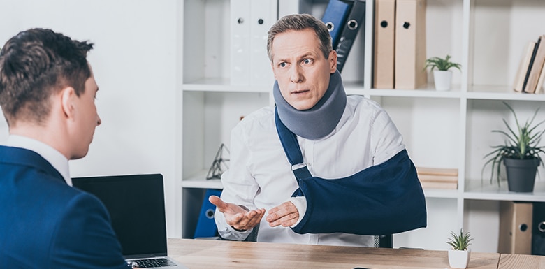 A man wearing a neck brace and arm sling sits at a desk, talking to another man in a suit who is using a laptop. The setting appears to be an office with shelves and potted plants in the background.