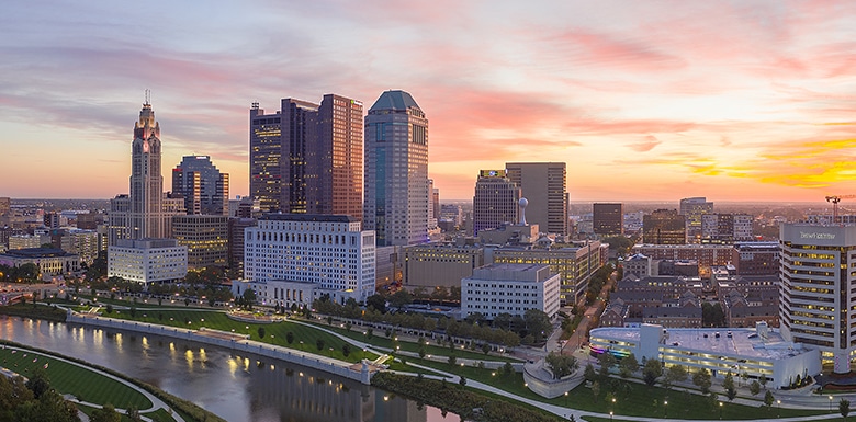 A city skyline at sunset with tall buildings, a river running through the foreground, and a colorful sky with pink and orange clouds.