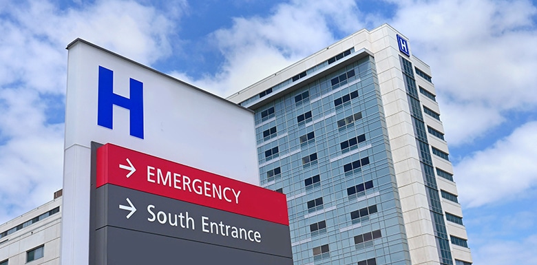 A hospital building with a large blue H sign. In the foreground, a directional sign points to EMERGENCY and South Entrance against a background of partly cloudy sky.