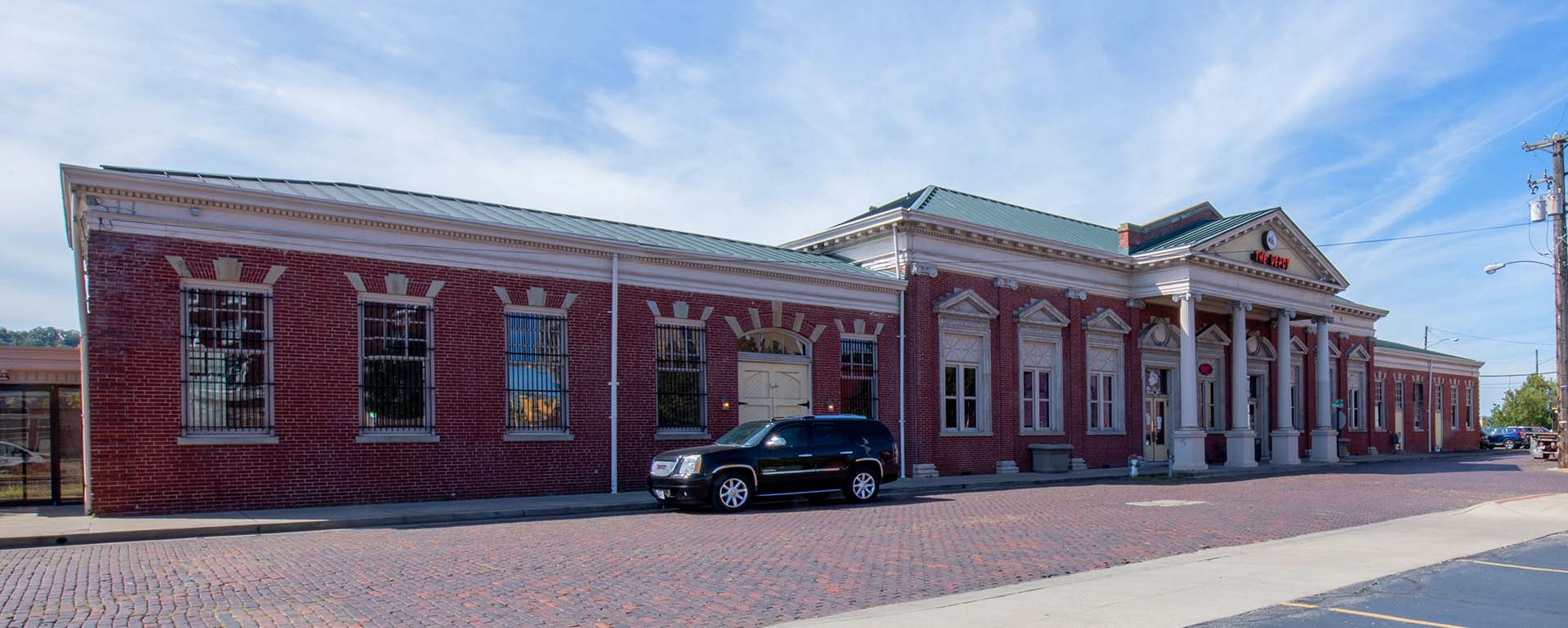 A historic red brick building with white trim and columns, featuring large windows and a green metal roof. A black SUV is parked in front on a cobblestone street under a blue sky.
