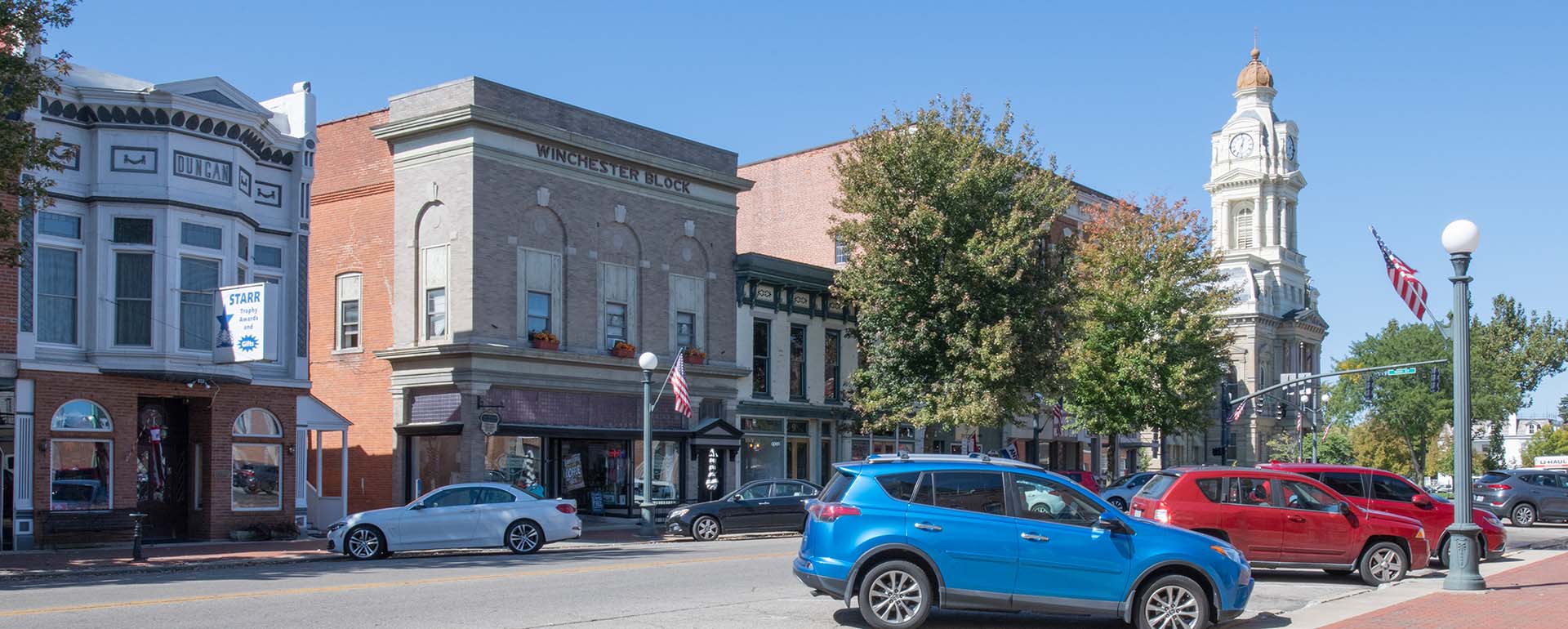 A small town main street with historic brick buildings, shops, and a tall clock tower. Cars are parked along the street under a clear blue sky, and trees line the sidewalk.