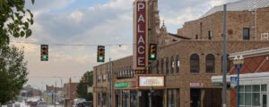 A street view of a historic brick theater building with a large vertical PALACE sign and a marquee reading The Music Man, Greham. Traffic lights hang above, and cars are parked along the road under a partly cloudy sky.