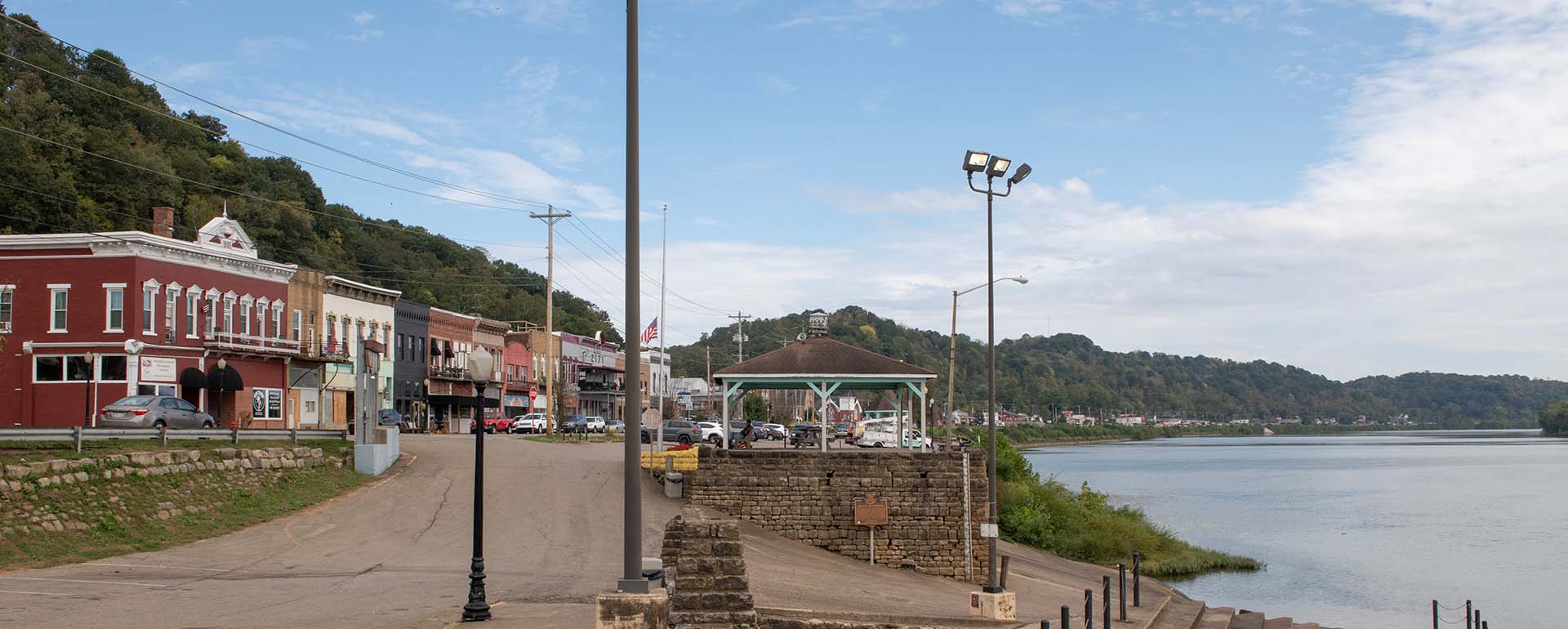 A small riverside town features historic brick buildings, a gazebo, lampposts, and a tree-covered hill. Cars are parked along the street, and the calm river runs alongside the scene under a partly cloudy sky.