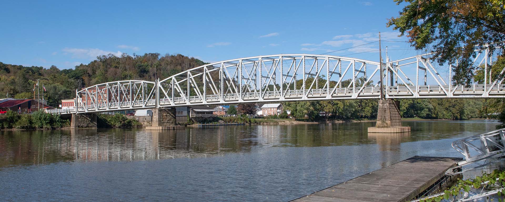 A white truss bridge spans a calm river, supported by stone piers. Trees and a few buildings are visible on both riverbanks under a clear blue sky.