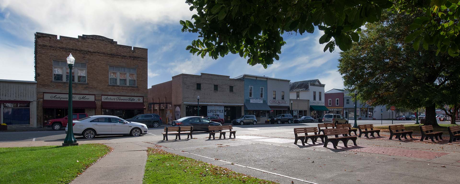 A small town main street with historic brick buildings, parked cars, wooden benches, and trees under a partly cloudy sky on a quiet day.