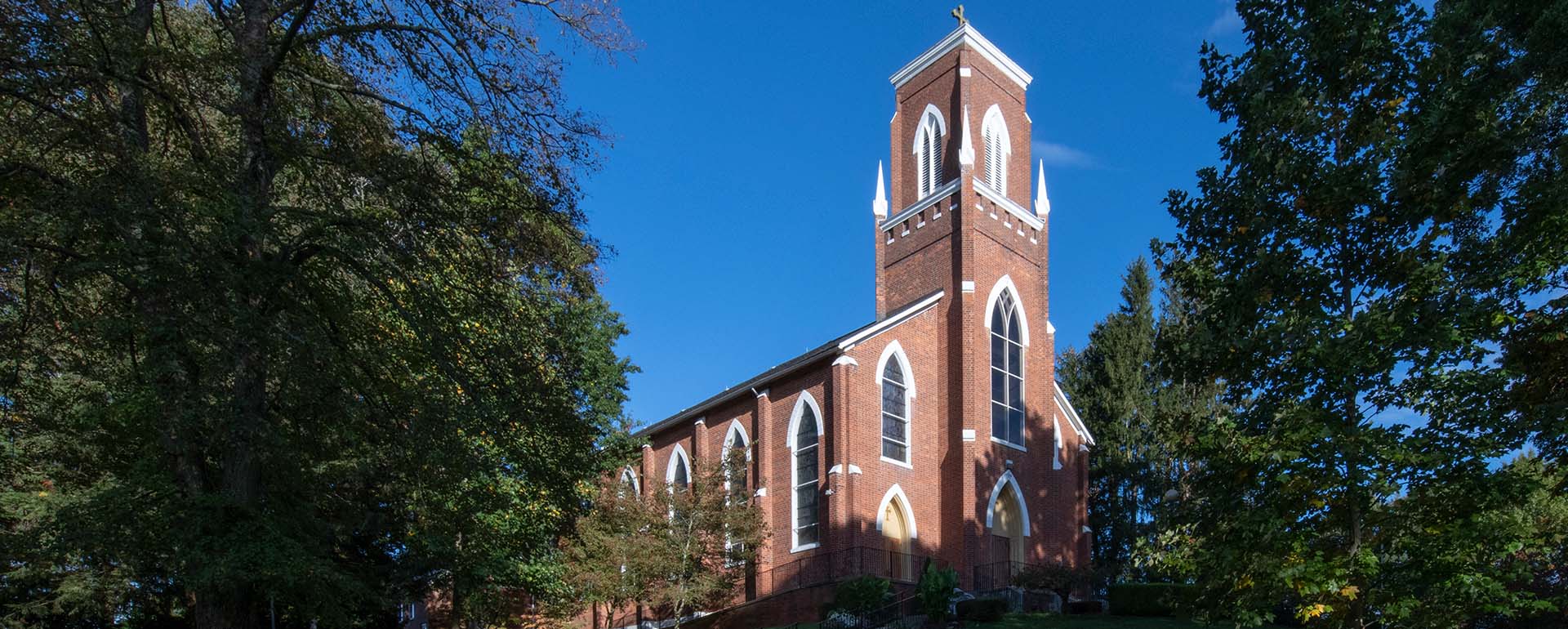 A red brick church with tall arched windows and a central tower topped by a cross, surrounded by green trees under a clear blue sky.