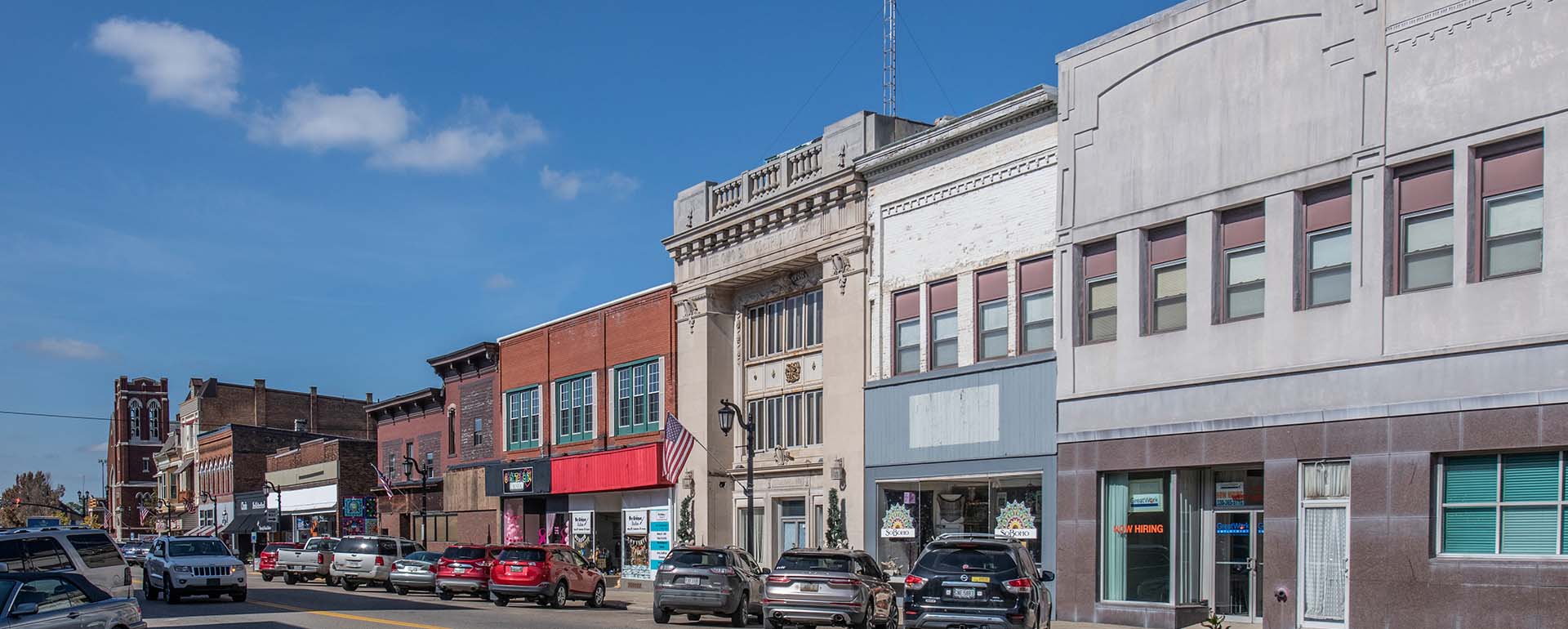 A row of historic downtown buildings lines a sunny street with parked cars, large windows, and various storefronts; blue sky and a few clouds are visible above.