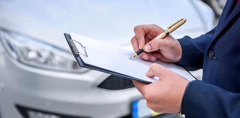 A person in a suit is writing on a clipboard with a pen, standing in front of a silver car.