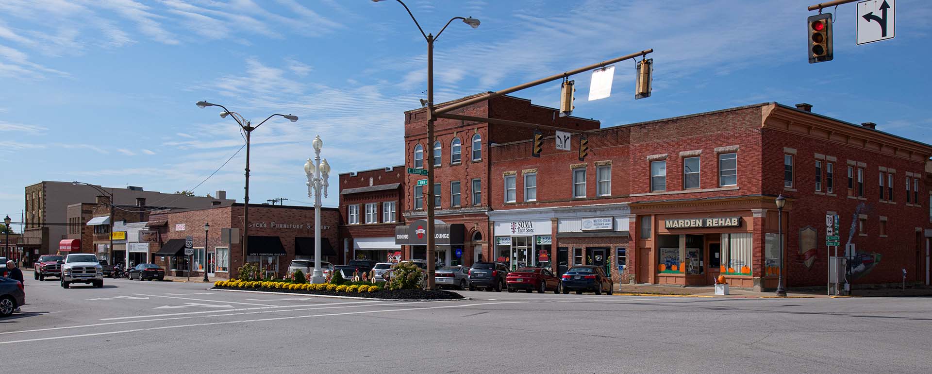 Small-town street corner with historic brick buildings, various storefronts, parked cars, and traffic lights. The sky is clear and blue, and flowers are planted in a median at the intersection.