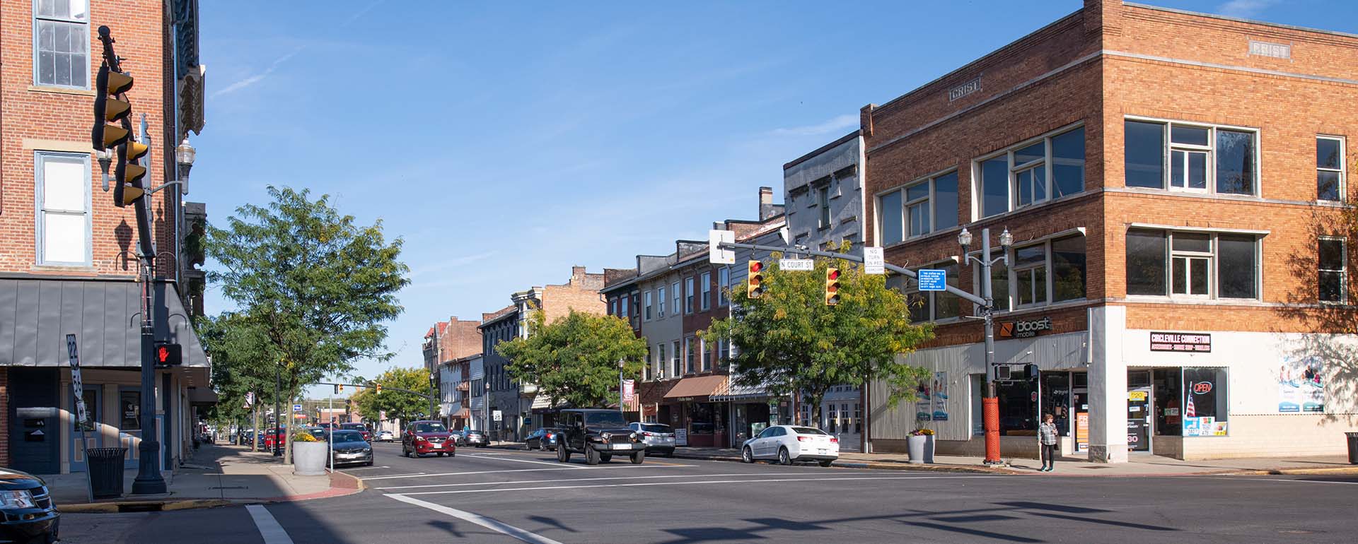 A sunny street scene in a small town shows cars driving and parked along the road, brick buildings with shops and businesses, trees lining the sidewalk, and a traffic light at an intersection.