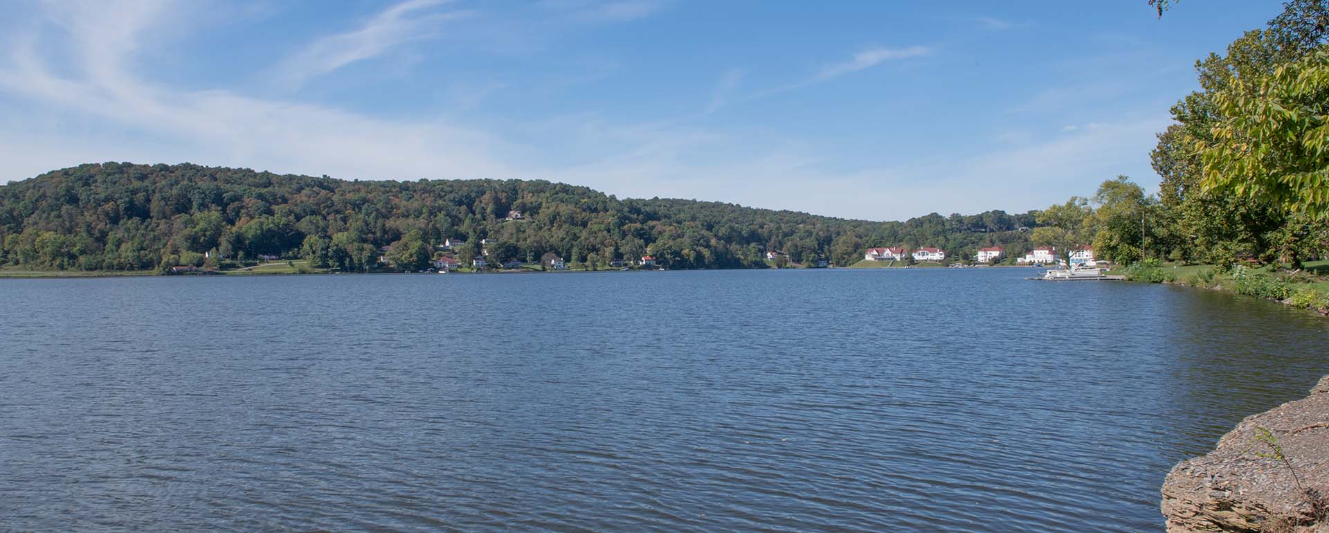 A wide lake with gentle ripples, bordered by a forested hillside and several lakeside houses under a blue sky with wispy clouds, viewed from the rocky shoreline.