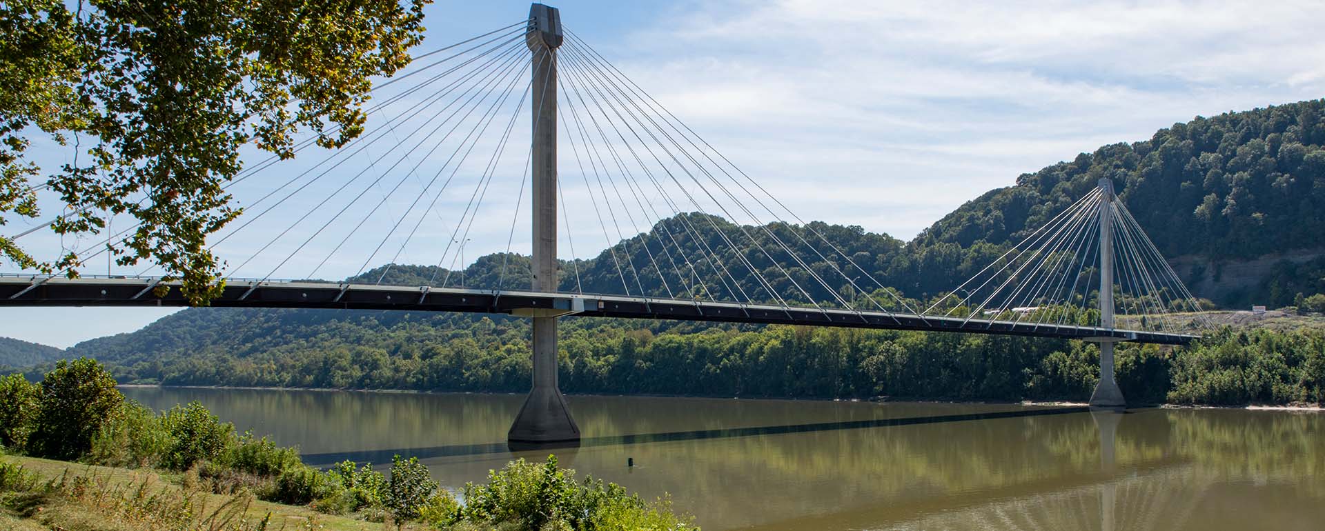 A cable-stayed bridge spans over a calm river, surrounded by green trees and hills under a partly cloudy sky.