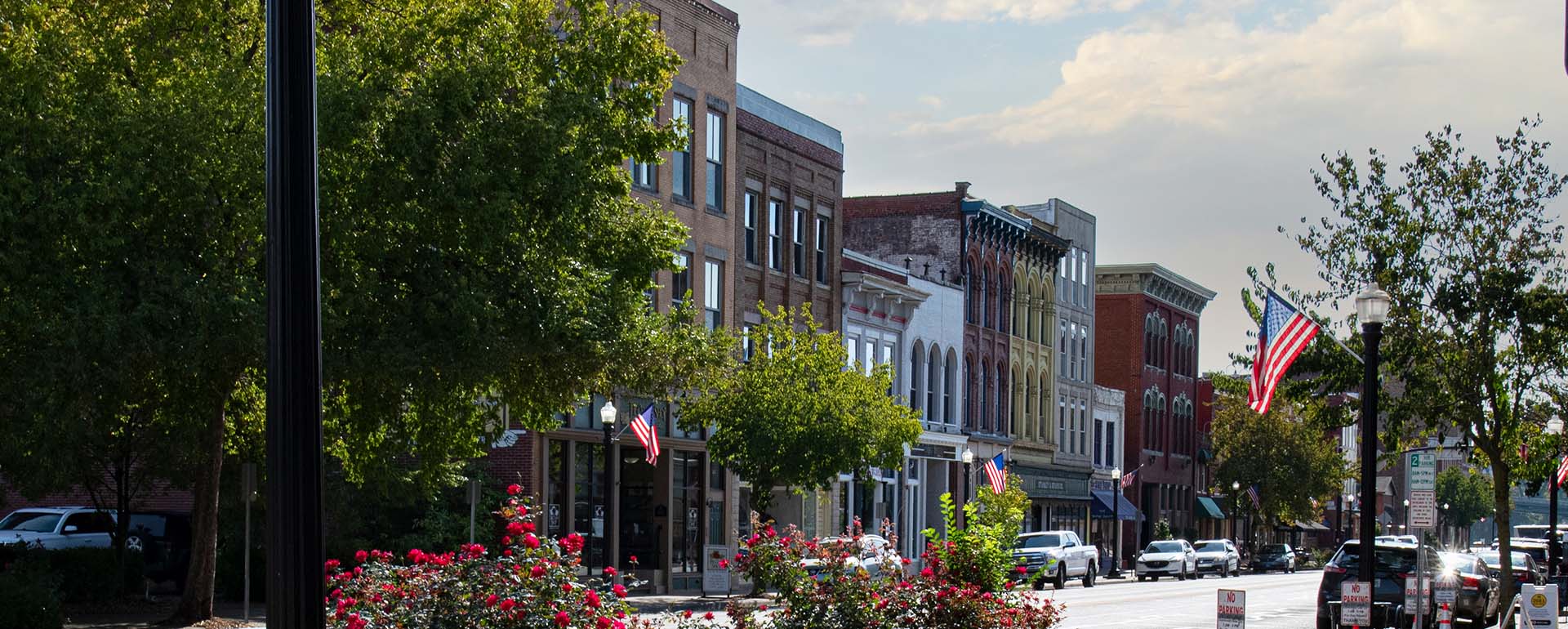 A small town main street with historic brick buildings, storefronts, American flags, blooming flowers, green trees, and parked cars on both sides under a partly cloudy sky.