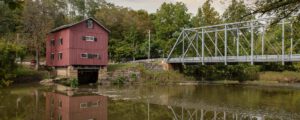 A red wooden mill sits on the edge of a river beside a silver metal truss bridge, surrounded by green trees and reflected in the calm water below.