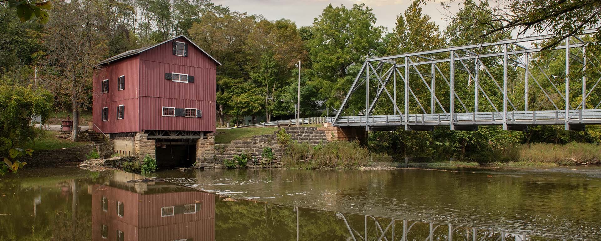 A red wooden mill sits on the edge of a river beside a silver metal truss bridge, surrounded by green trees and reflected in the calm water below.