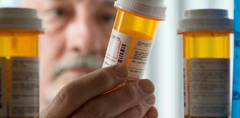 A person holds a prescription pill bottle and examines the label, with two more orange prescription bottles visible in the foreground. The persons face is out of focus in the background.