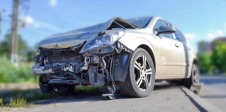 A silver car with severe front-end damage is parked on the side of a road. The hood is crumpled and parts of the bumper and engine are exposed, indicating a recent collision. The background is blurred.