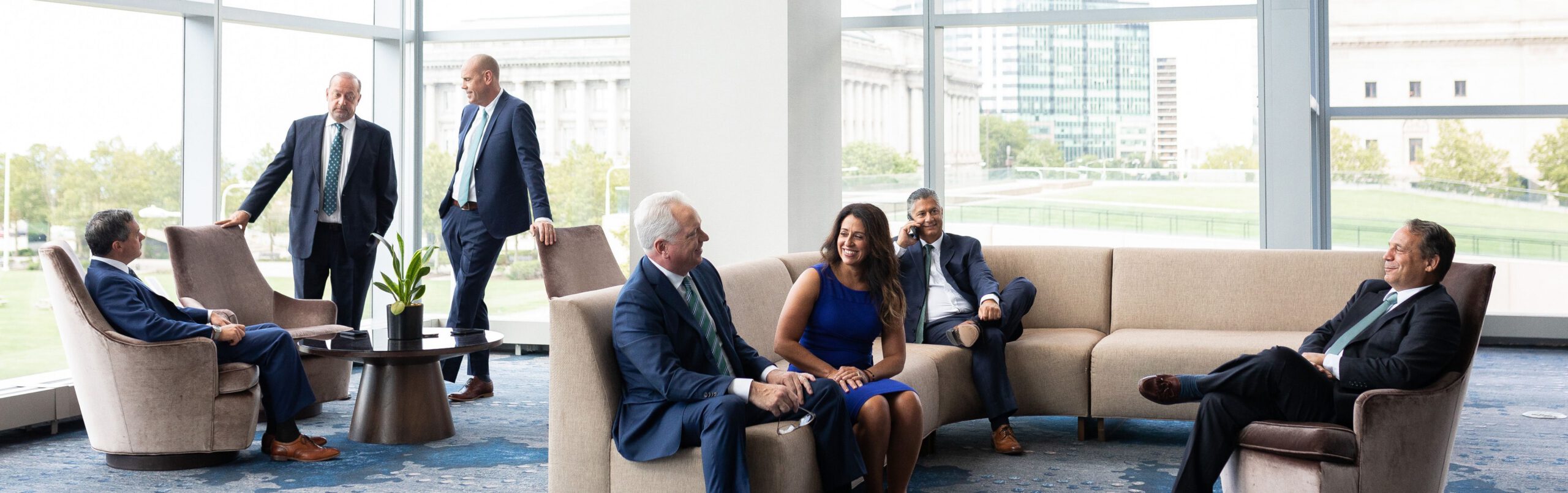 A group of six people in business attire sit and stand in a modern office lounge with large windows, smiling and conversing. The room has beige sofas, chairs, and a plant on a table. City buildings are visible outside.
