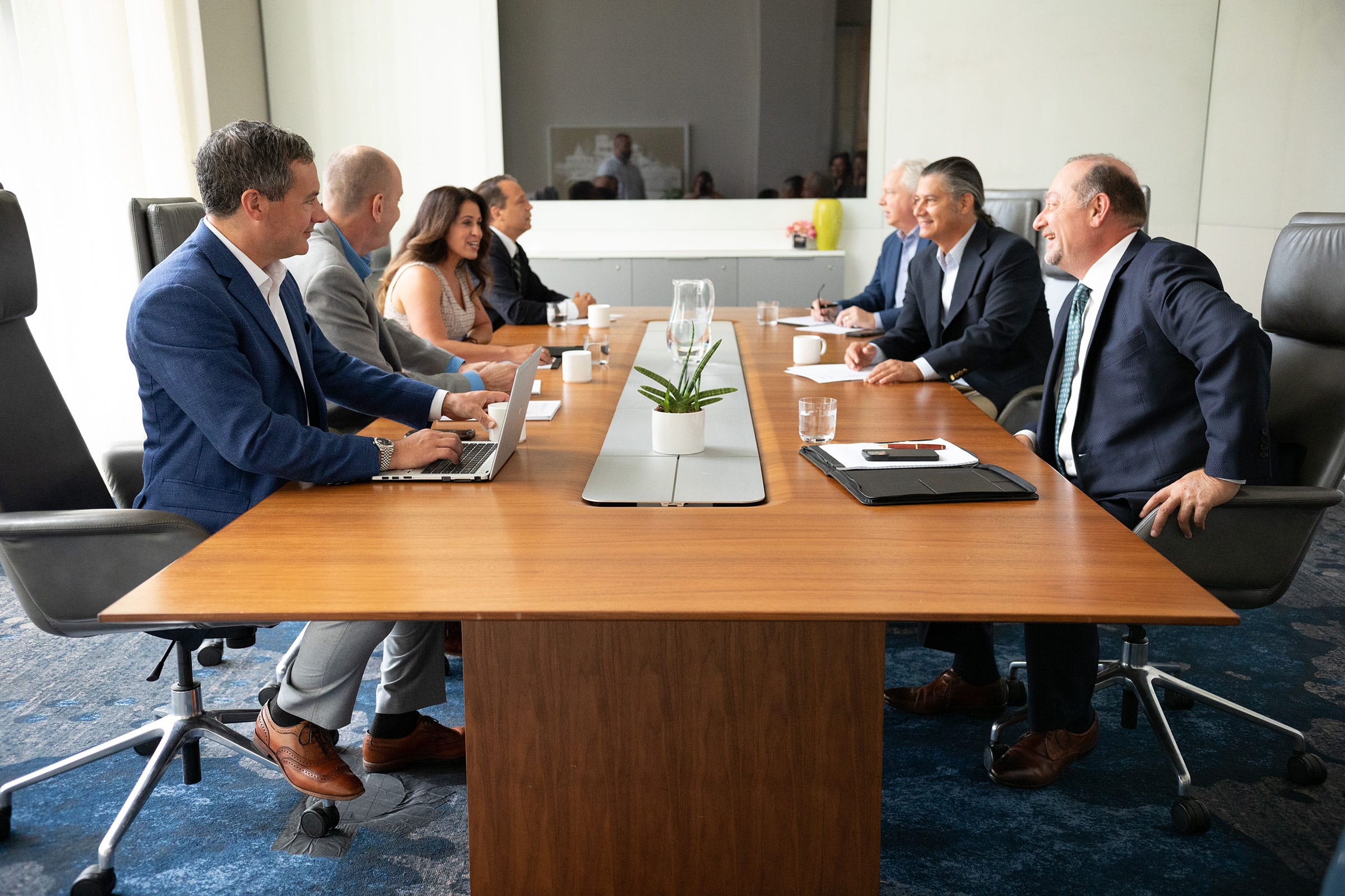 A group of business professionals, both men and women, sit around a conference table engaged in a meeting. They appear to be discussing something and smiling. There are laptops, papers, and a plant on the table.