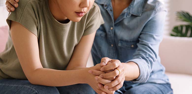 Two women sit close together; one looks sad and is leaning forward with clasped hands, while the other gently comforts her by placing a hand on her back and holding her hand supportively.