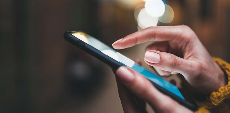 A close-up of a person’s hands using a smartphone, with one hand holding the device and the other hand tapping the screen. The background is blurred with bokeh lights.