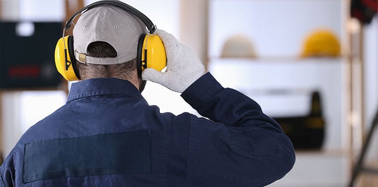 A person wearing a blue work jacket, gray cap, and yellow ear protection stands indoors, seen from behind. They are also wearing a white glove and appear to be in a workshop or industrial setting.