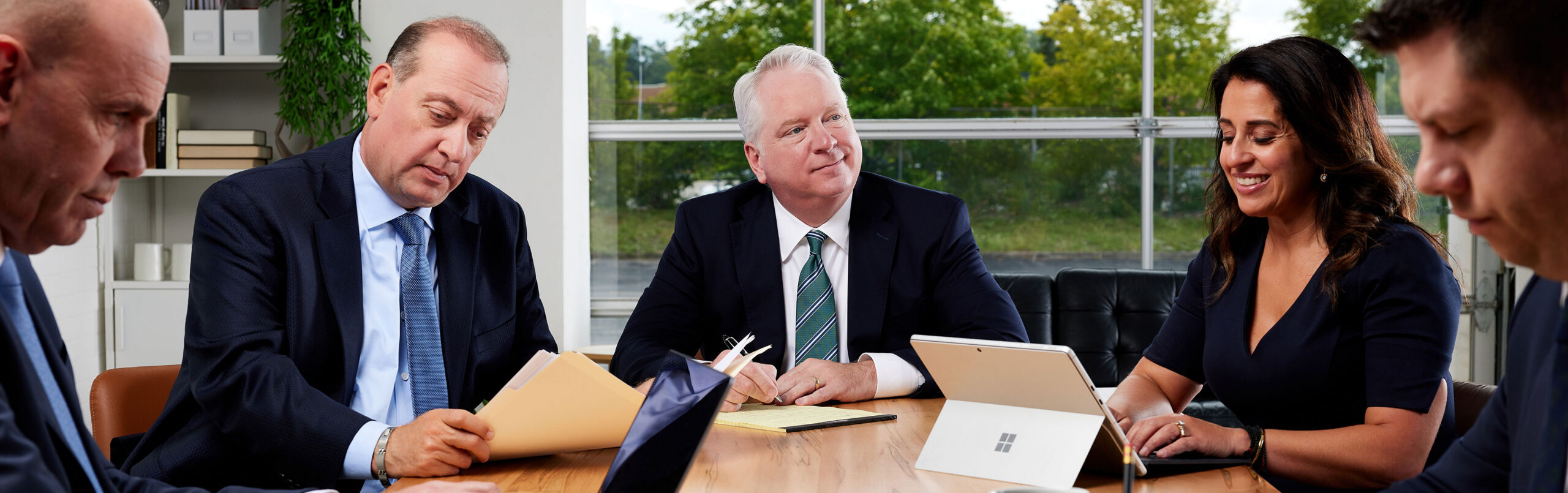 Five professionally dressed people sit around a conference table, using laptops and reviewing documents. Large windows and greenery are visible in the background, suggesting a modern office setting.