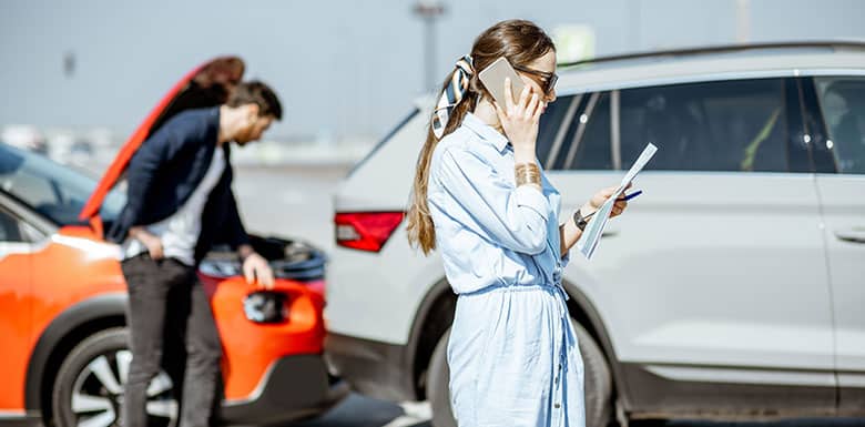 A woman stands beside a silver SUV, talking on her phone and holding paperwork. In the background, a man inspects the engine of an orange car with its hood open, suggesting a possible car accident or breakdown.