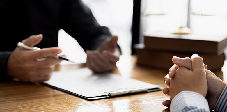 Two people sit at a wooden table having a discussion; one gestures with a pen near a clipboard, while the other has hands clasped. Books and scales of justice are visible in the background.