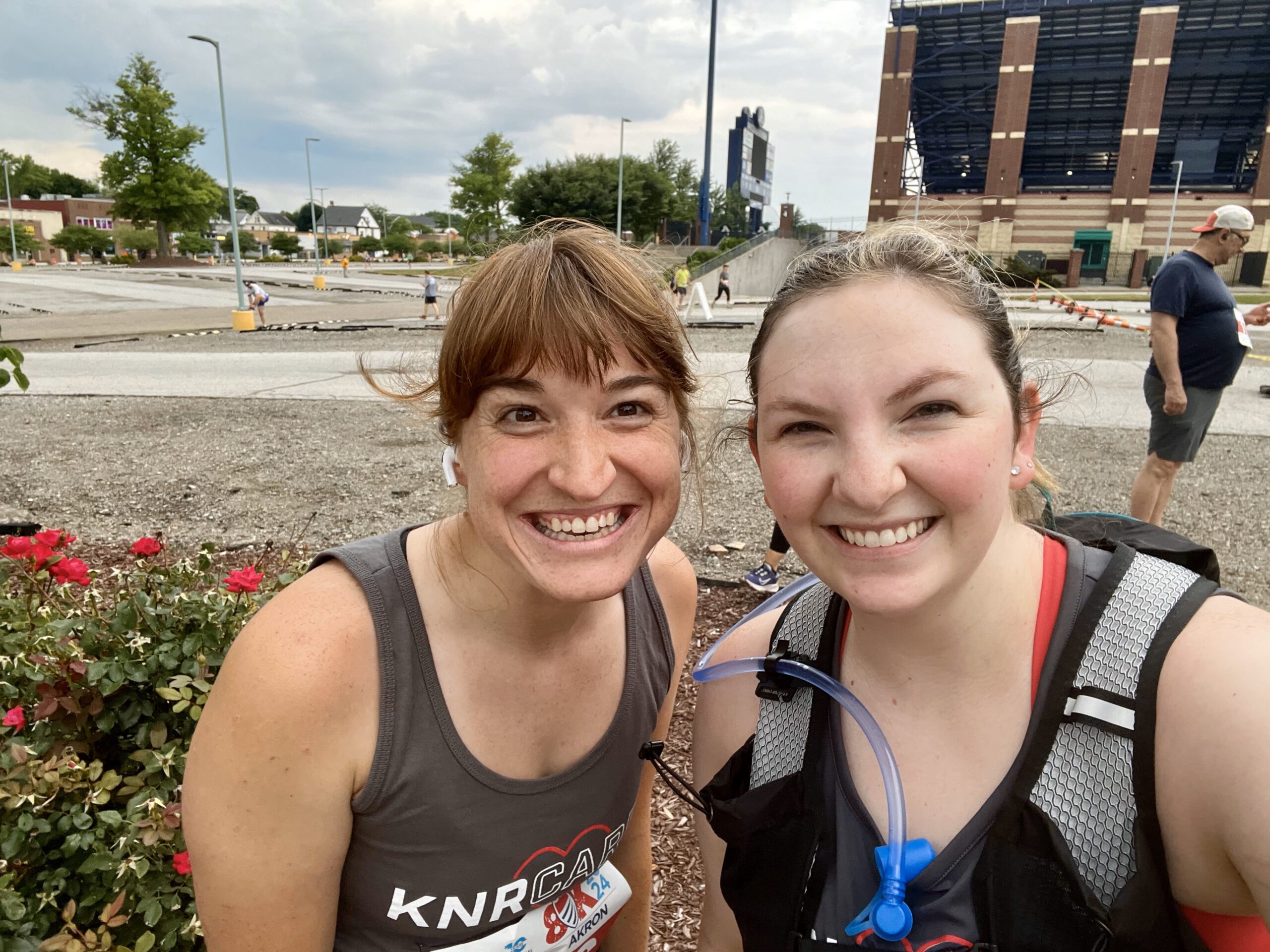 Two women smiling for a selfie outdoors, both wearing athletic gear. One has a hydration pack, the other wears earbuds. They stand near red flowers with a stadium and parking lot in the background.