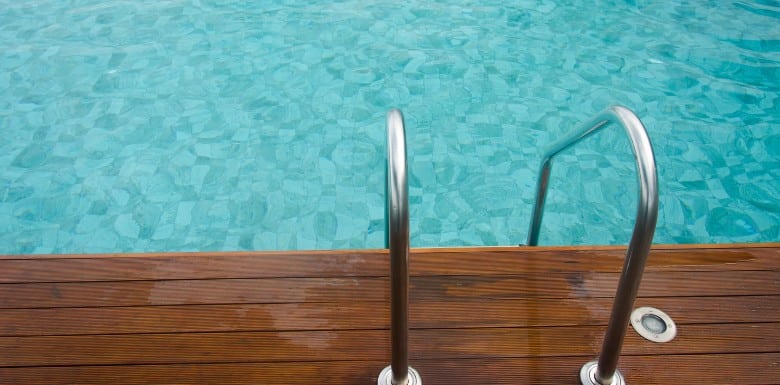 Metal pool ladder attached to a wooden deck, leading into a clear blue swimming pool with tiled floor visible under the water.
