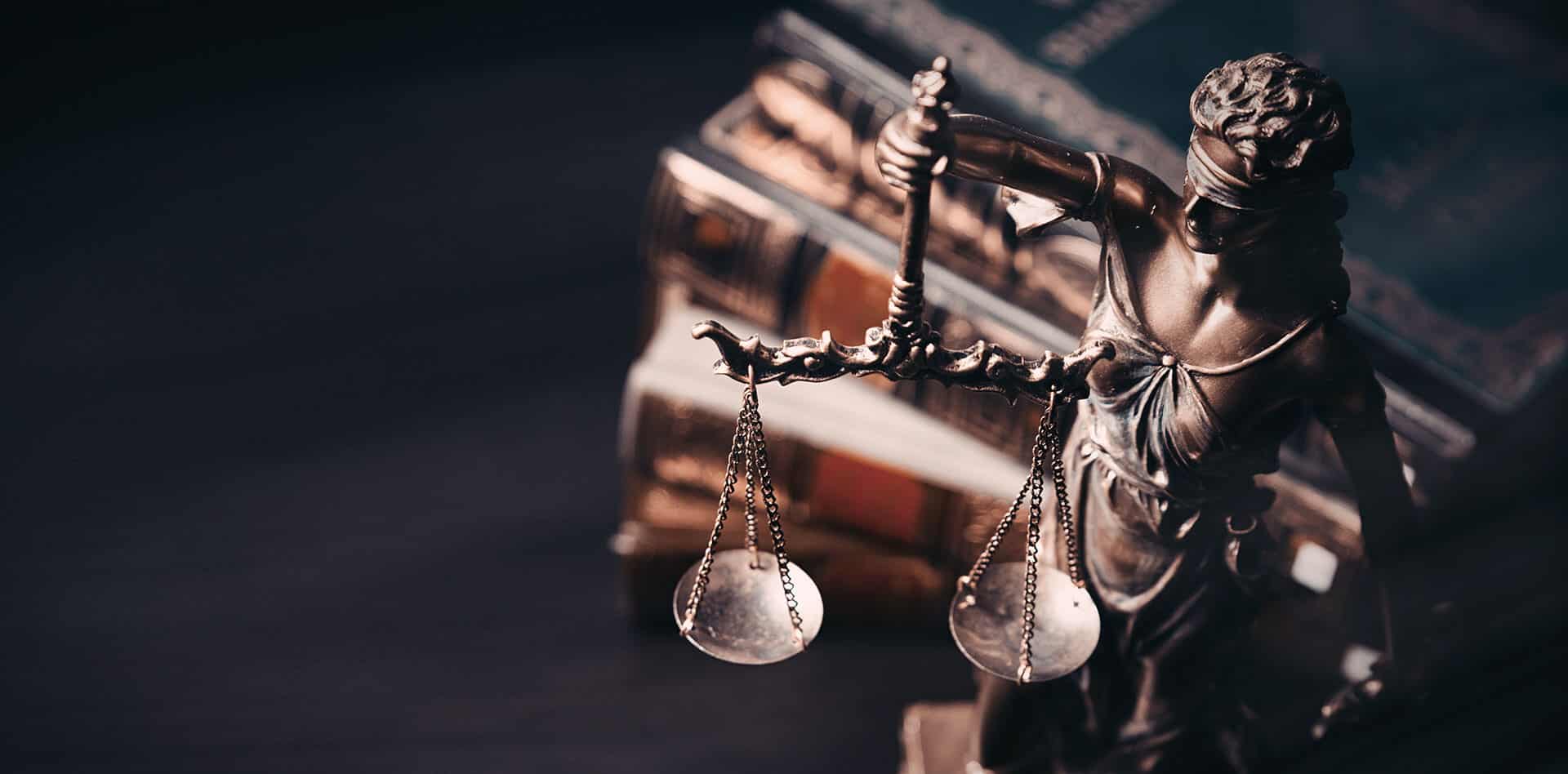 A close-up of a bronze Lady Justice statue holding balanced scales, with stacked law books in the background. The image symbolizes justice, law, and legal proceedings.