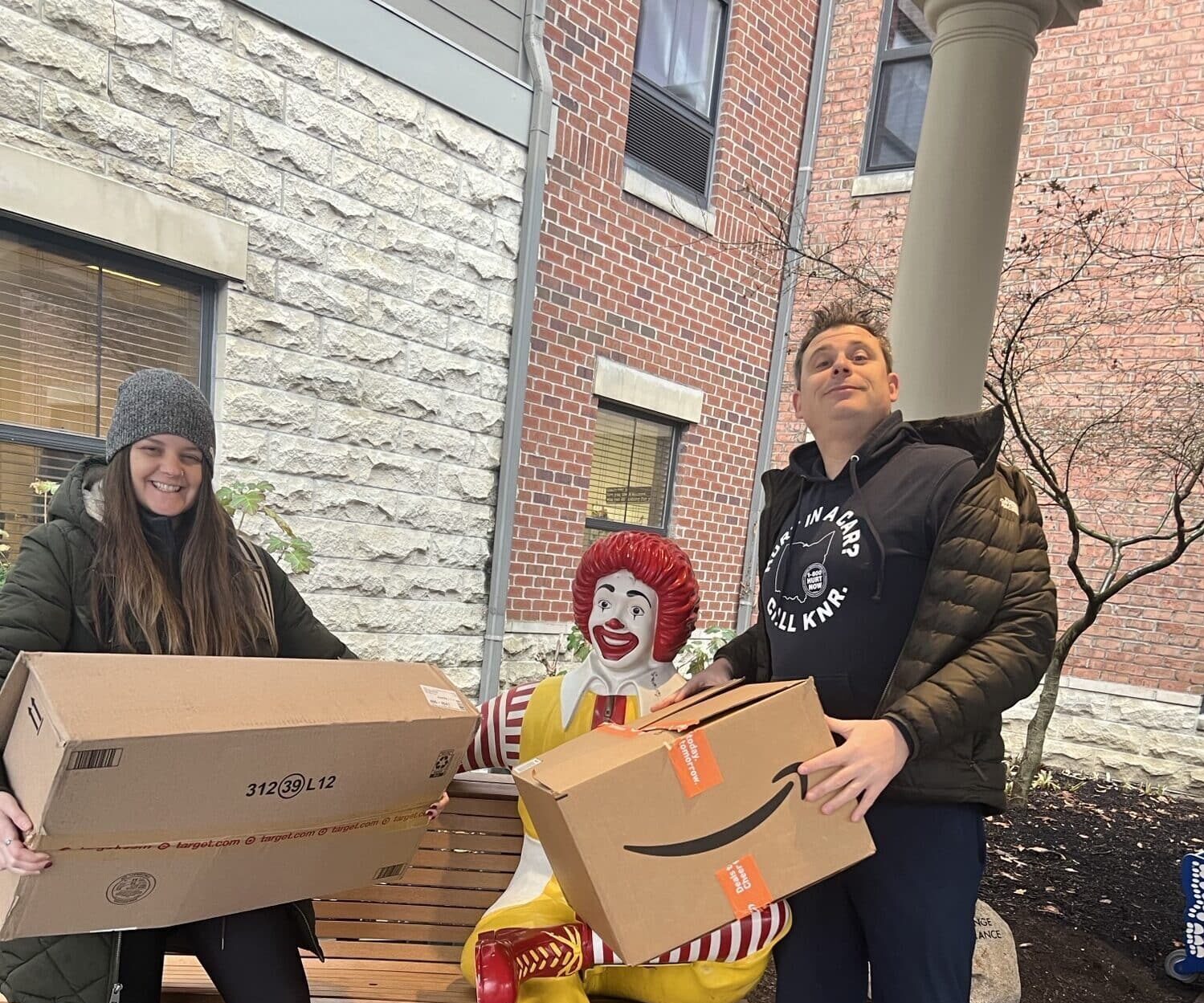 A woman and a man smile while holding large Amazon boxes next to a Ronald McDonald statue on a bench outside a brick building.