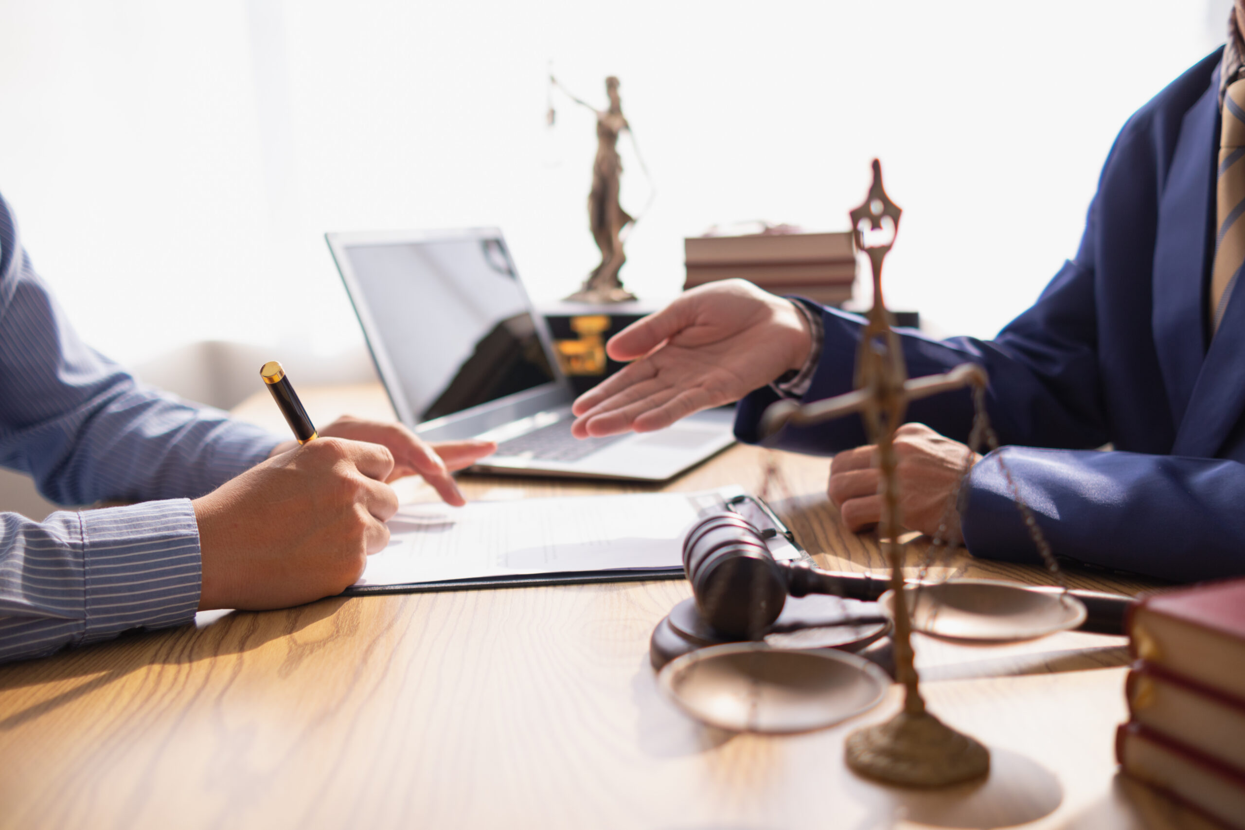 Two people sit at a desk with legal documents, a gavel, and scales of justice. One person is signing papers while the other gestures, suggesting a legal consultation or agreement. A laptop and law books are in the background.