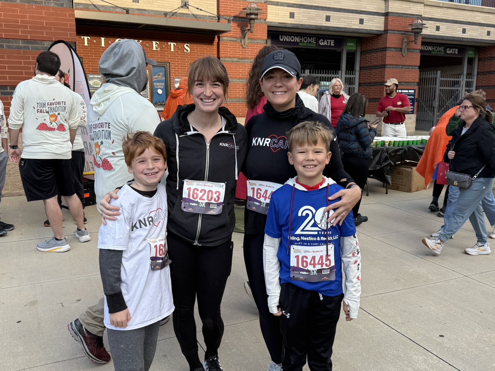 Two women and two boys smile at a race event, wearing running bibs and athletic wear. Other participants and vendors are visible in the background near a brick building.