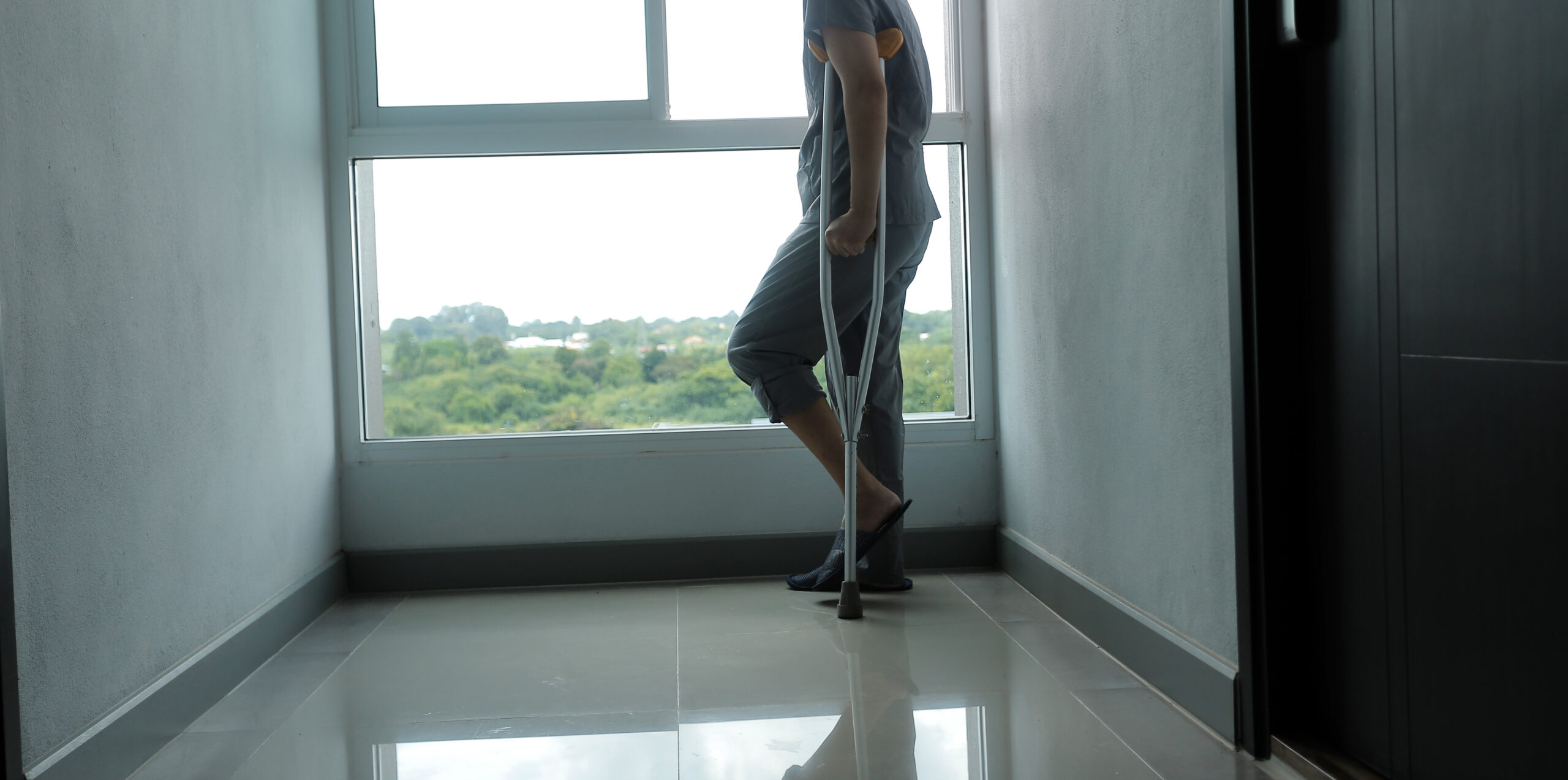 A person with cropped hair stands near a large window in a hallway, using crutches. Only the lower half of their body is visible. Greenery and trees can be seen outside through the window.