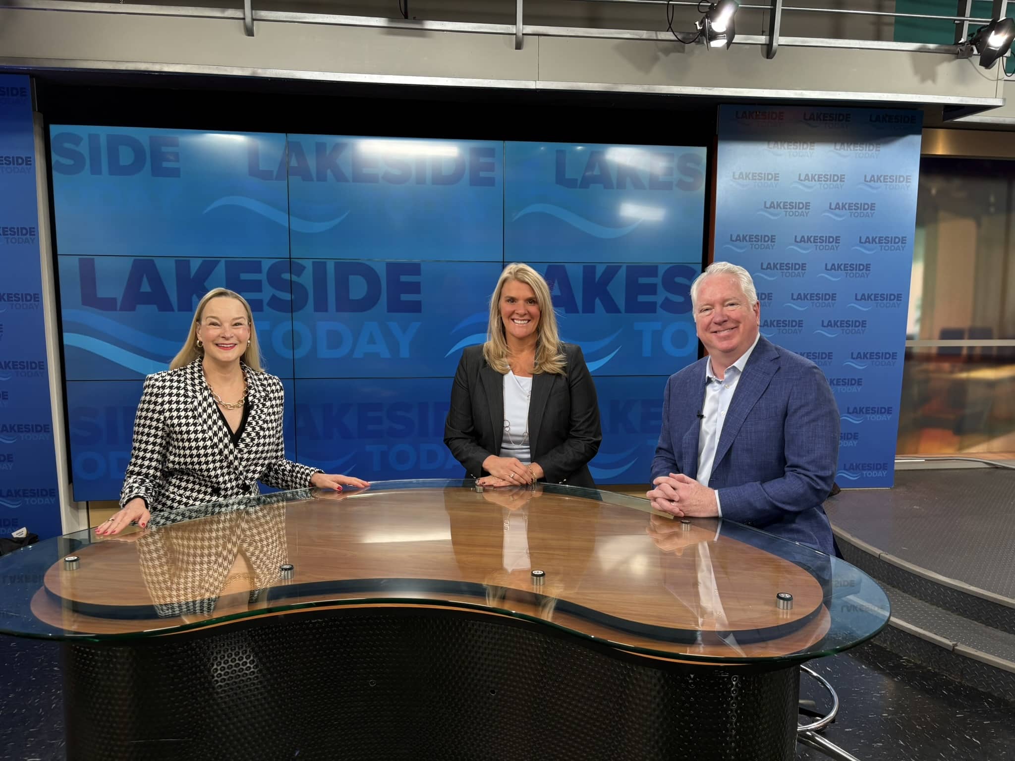 Three people, two women and one man, sit smiling at a curved news desk in a brightly lit TV studio with LAKESIDE TODAY and blue backgrounds behind them.